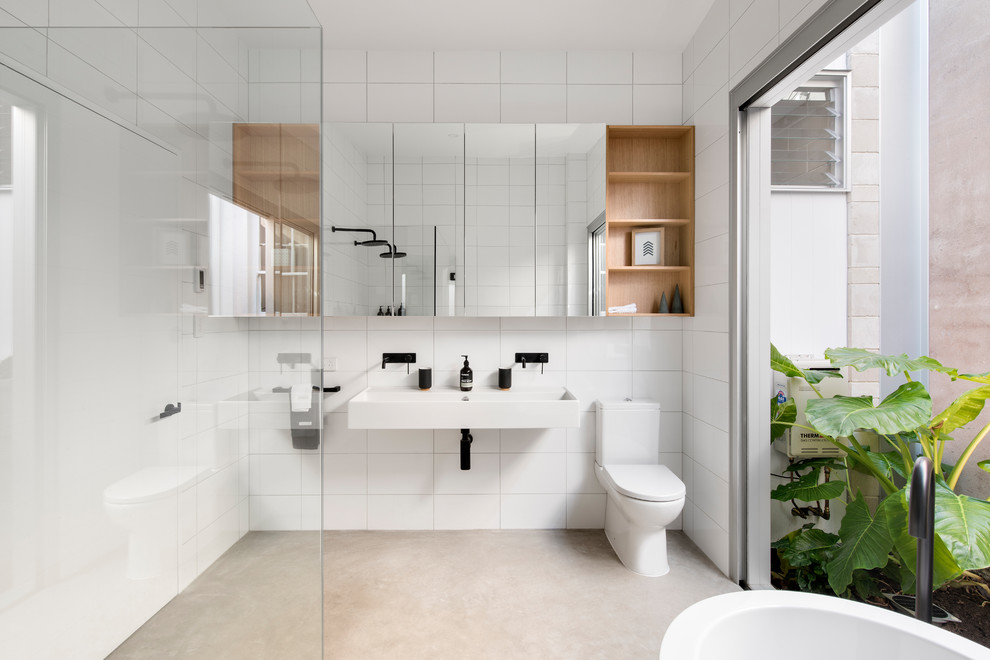 This is an example of a medium sized contemporary ensuite bathroom in Adelaide with freestanding cabinets, light wood cabinets, a freestanding bath, a double shower, a wall mounted toilet, white tiles, ceramic tiles, white walls, concrete flooring and a wall-mounted sink.