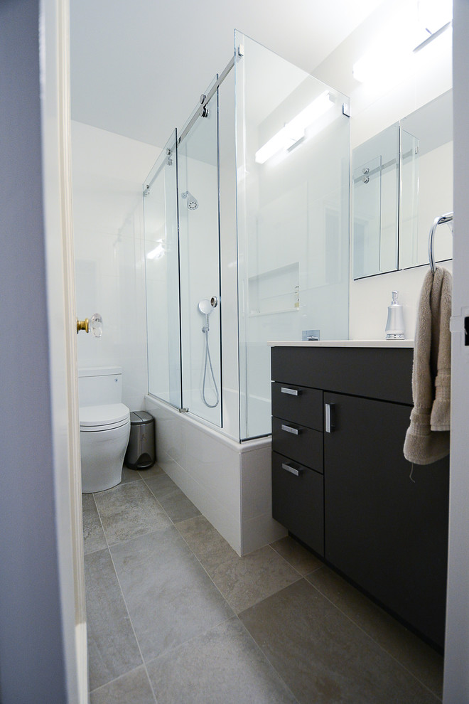 Пример оригинального дизайна: главная ванная комната среднего размера в стиле модернизм с плоскими фасадами, черными фасадами, угловой ванной, душем над ванной, унитазом-моноблоком, белой плиткой, белыми стенами, полом из керамогранита, подвесной раковиной, серым полом, душем с раздвижными дверями и белой столешницей