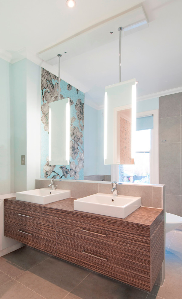 Photo of a large contemporary ensuite wet room bathroom in London with flat-panel cabinets, medium wood cabinets, wooden worktops, grey floors, brown worktops, double sinks, a floating vanity unit, a freestanding bath, a wall mounted toilet, grey tiles, mosaic tiles, blue walls, porcelain flooring, a console sink and an open shower.
