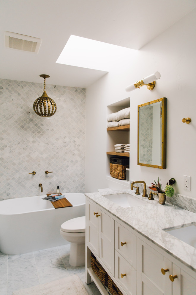 Skandinavisk inredning av ett grå grått badrum med dusch, med skåp i shakerstil, vita skåp, ett fristående badkar, en toalettstol med separat cisternkåpa, vita väggar, ett undermonterad handfat och grått golv