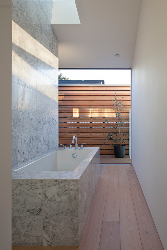 Aménagement d'une salle de bain principale moderne de taille moyenne avec un placard à porte plane, des portes de placard blanches, un plan de toilette en marbre, une baignoire encastrée, un carrelage de pierre et un sol en bois brun.