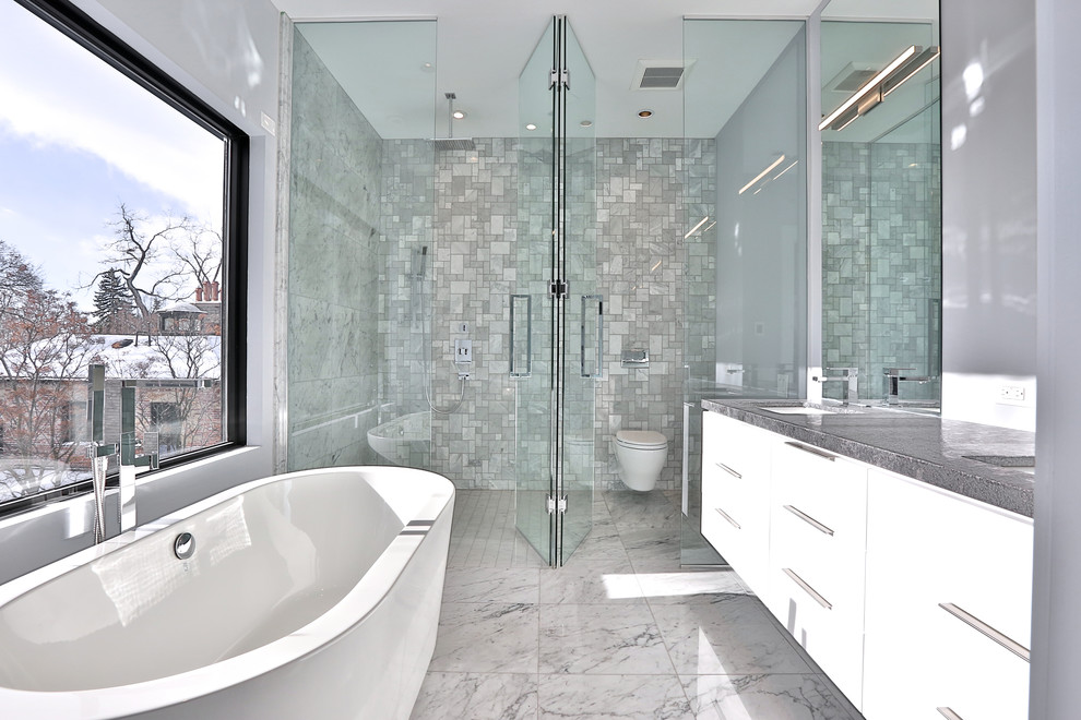 На фото: большая главная ванная комната в стиле модернизм с плоскими фасадами, белыми фасадами, отдельно стоящей ванной, душем без бортиков, инсталляцией, серой плиткой, мраморной плиткой, серыми стенами, мраморным полом, врезной раковиной, столешницей из гранита, разноцветным полом и душем с распашными дверями