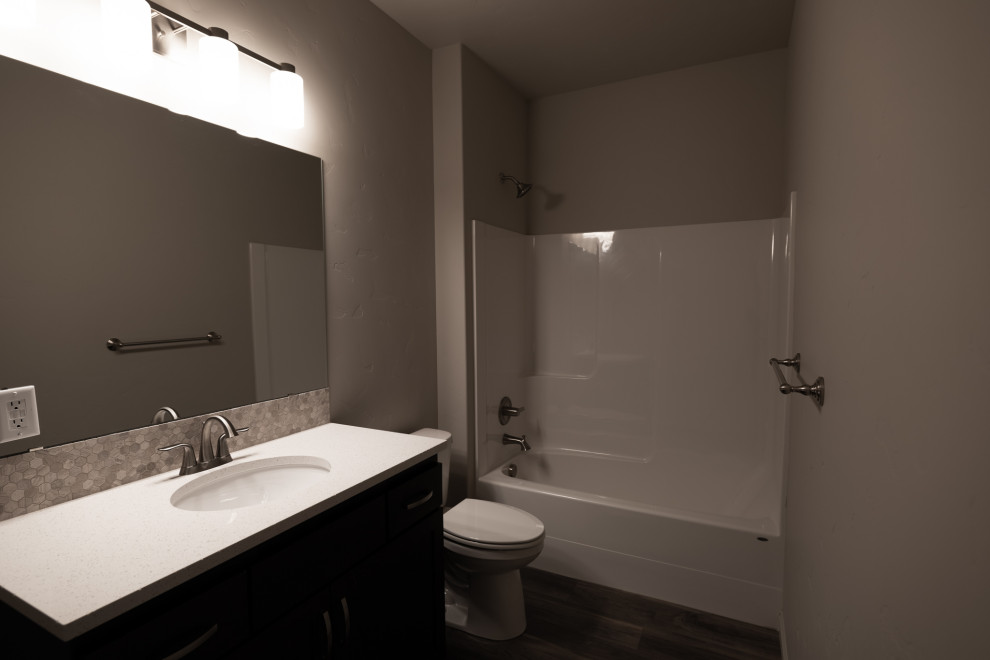 Идея дизайна: маленькая ванная комната в стиле кантри с плоскими фасадами, темными деревянными фасадами, ванной в нише, душем над ванной, раздельным унитазом, разноцветной плиткой, керамической плиткой, серыми стенами, полом из винила, врезной раковиной, столешницей из искусственного кварца, коричневым полом, шторкой для ванной, белой столешницей, тумбой под одну раковину, встроенной тумбой и фартуком для на участке и в саду