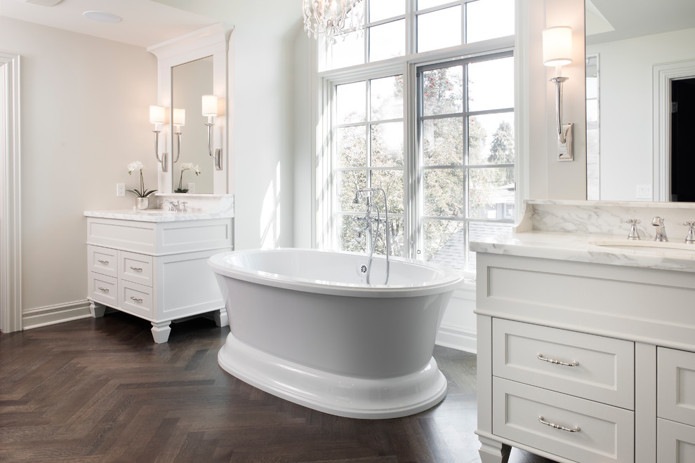 На фото: большая главная ванная комната в классическом стиле с фасадами в стиле шейкер, белыми фасадами, столешницей из искусственного камня, отдельно стоящей ванной и паркетным полом среднего тона