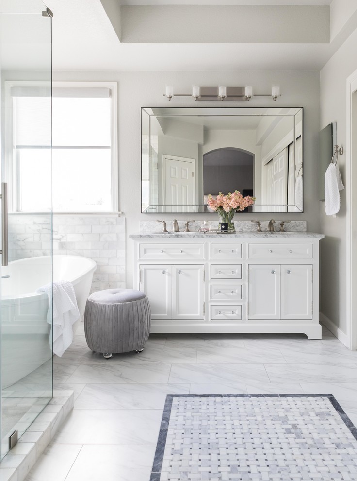 Cette image montre une salle de bain principale traditionnelle de taille moyenne avec un placard à porte shaker, des portes de placard blanches, une baignoire indépendante, un carrelage blanc, du carrelage en marbre, un mur gris, un sol en marbre, un lavabo encastré, un plan de toilette en marbre, un sol blanc, une cabine de douche à porte battante et un plan de toilette blanc.
