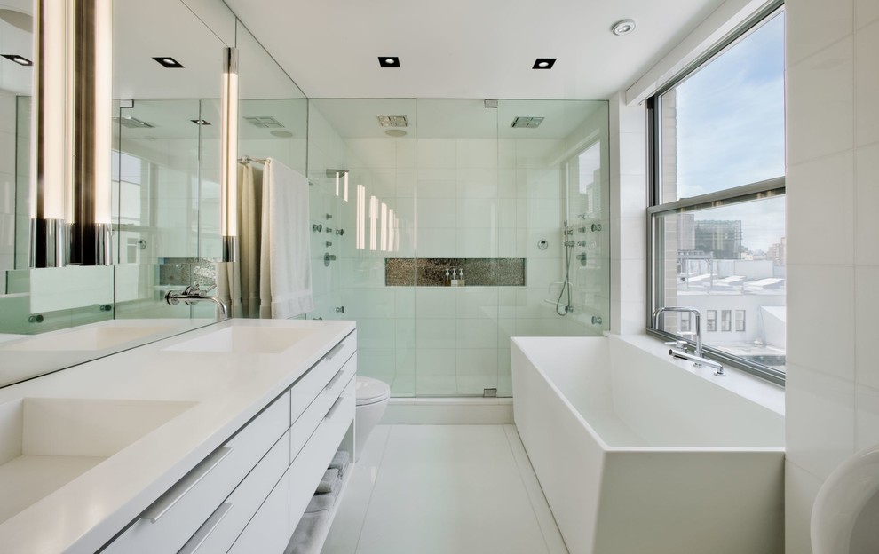 This is an example of a medium sized modern ensuite bathroom in New York with flat-panel cabinets, white cabinets, a freestanding bath, an alcove shower, a one-piece toilet, white tiles, porcelain tiles, white walls, porcelain flooring, an integrated sink, engineered stone worktops, white floors, a hinged door and white worktops.