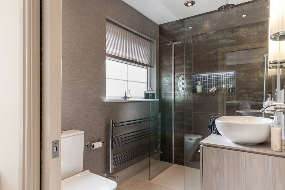 Modern inredning av ett en-suite badrum, med en toalettstol med hel cisternkåpa, svart kakel, släta luckor, grå skåp, våtrum, bruna väggar, ett fristående handfat, beiget golv och med dusch som är öppen