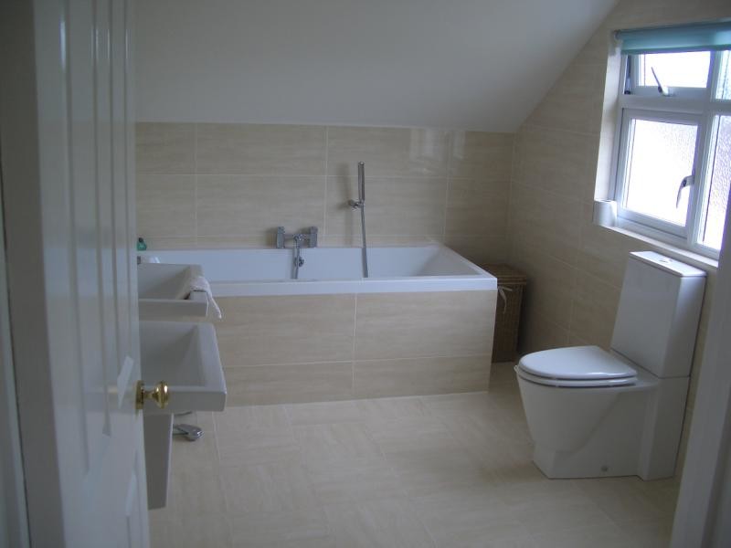 Photo of a medium sized modern ensuite bathroom in Gloucestershire with a wall-mounted sink, a built-in bath, a one-piece toilet, beige tiles, beige walls and ceramic flooring.