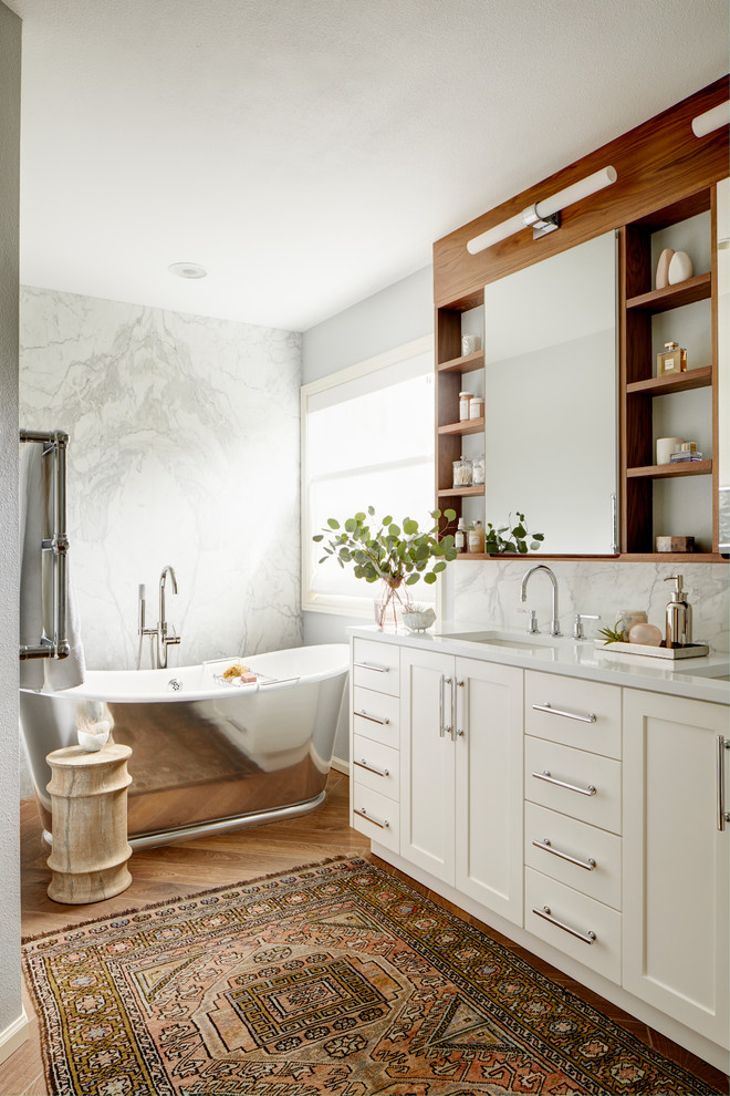 Idées déco pour une salle de bain principale classique avec un placard à porte shaker, des portes de placard blanches, une baignoire indépendante, un carrelage gris, un mur gris, un sol en bois brun, un sol marron et un plan de toilette blanc.
