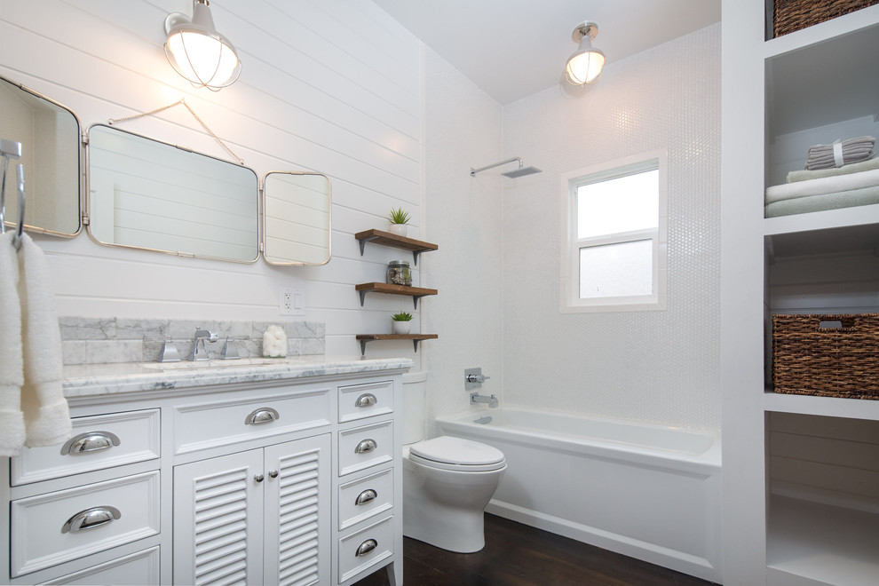 Minimalistisk inredning av ett mellanstort badrum, med luckor med lamellpanel, marmorbänkskiva, ett badkar i en alkov, en dusch/badkar-kombination, en toalettstol med separat cisternkåpa, vit kakel, tunnelbanekakel, vita väggar, mörkt trägolv, vita skåp och ett undermonterad handfat