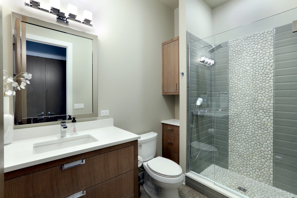 This is an example of a medium sized contemporary bathroom in Grand Rapids with flat-panel cabinets, dark wood cabinets, an alcove shower, a one-piece toilet, grey tiles, pebble tiles, grey walls, ceramic flooring, a submerged sink, engineered stone worktops, grey floors, a sliding door and white worktops.