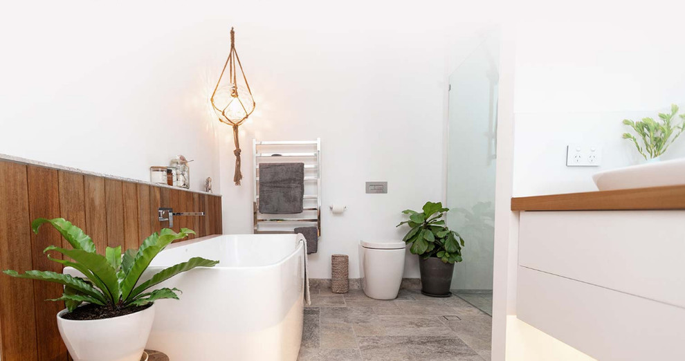Bild på ett stort funkis brun brunt en-suite badrum, med luckor med infälld panel, vita skåp, ett fristående badkar, en kantlös dusch, en vägghängd toalettstol, vit kakel, porslinskakel, vita väggar, travertin golv, ett fristående handfat, träbänkskiva, grått golv och med dusch som är öppen