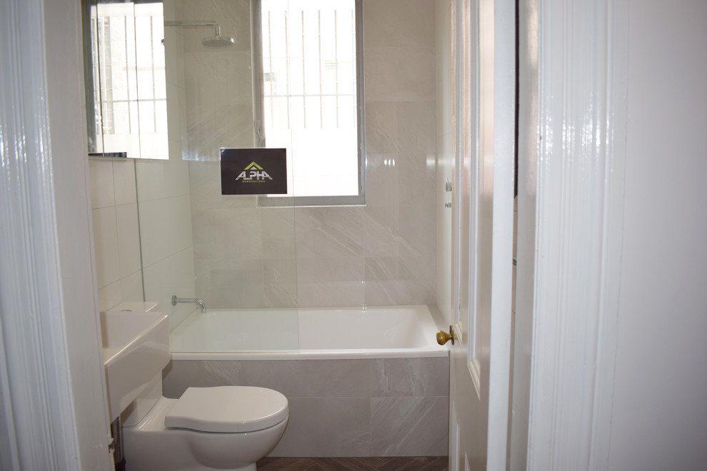 Foto på ett litet funkis en-suite badrum, med släta luckor, vita skåp, ett platsbyggt badkar, en dusch/badkar-kombination, en toalettstol med separat cisternkåpa, vit kakel, keramikplattor, vita väggar, klinkergolv i porslin, ett väggmonterat handfat, brunt golv och med dusch som är öppen