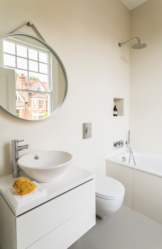 Design ideas for a medium sized contemporary ensuite bathroom in London with flat-panel cabinets, white cabinets, a built-in bath, a shower/bath combination, a wall mounted toilet, beige tiles, cement tiles, beige walls, cement flooring, a vessel sink, grey floors and white worktops.