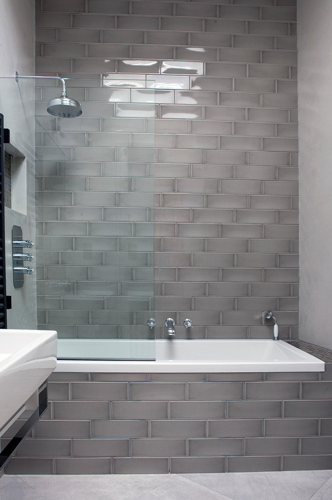Photo of a contemporary bathroom in London with a built-in bath, a shower/bath combination, a wall mounted toilet, grey tiles, ceramic tiles, grey walls, cement flooring, a wall-mounted sink, grey floors and a hinged door.