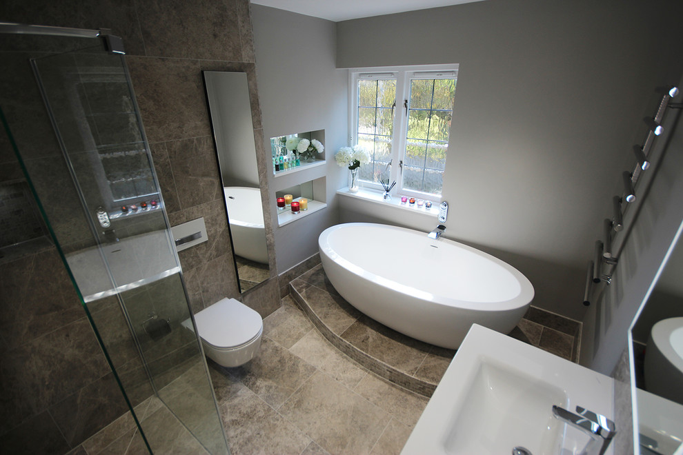 This is an example of a large contemporary ensuite wet room bathroom in Surrey with flat-panel cabinets, grey cabinets, a freestanding bath, a wall mounted toilet, grey tiles, stone tiles, grey walls, limestone flooring, a wall-mounted sink and grey floors.
