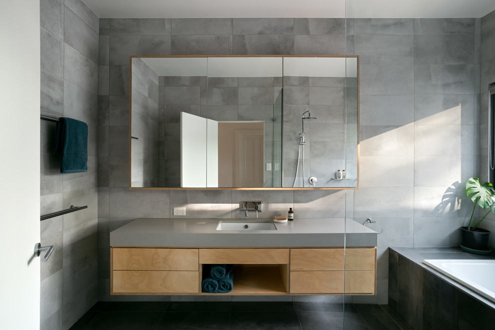 Bild på ett funkis grå grått badrum, med släta luckor, skåp i mellenmörkt trä, ett platsbyggt badkar, grå kakel, grå väggar, ett undermonterad handfat och svart golv
