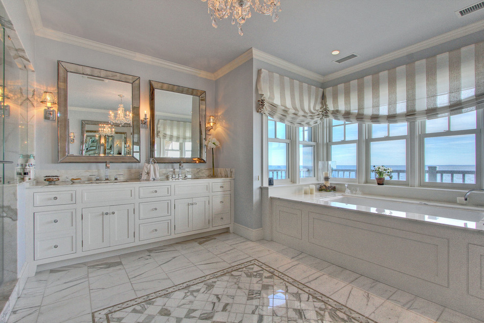 Cette photo montre une grande salle de bain principale bord de mer avec un lavabo intégré, un placard avec porte à panneau surélevé, des portes de placard blanches, un plan de toilette en marbre, une baignoire posée, un mur gris, un sol en marbre, une douche ouverte, un carrelage blanc et un carrelage de pierre.