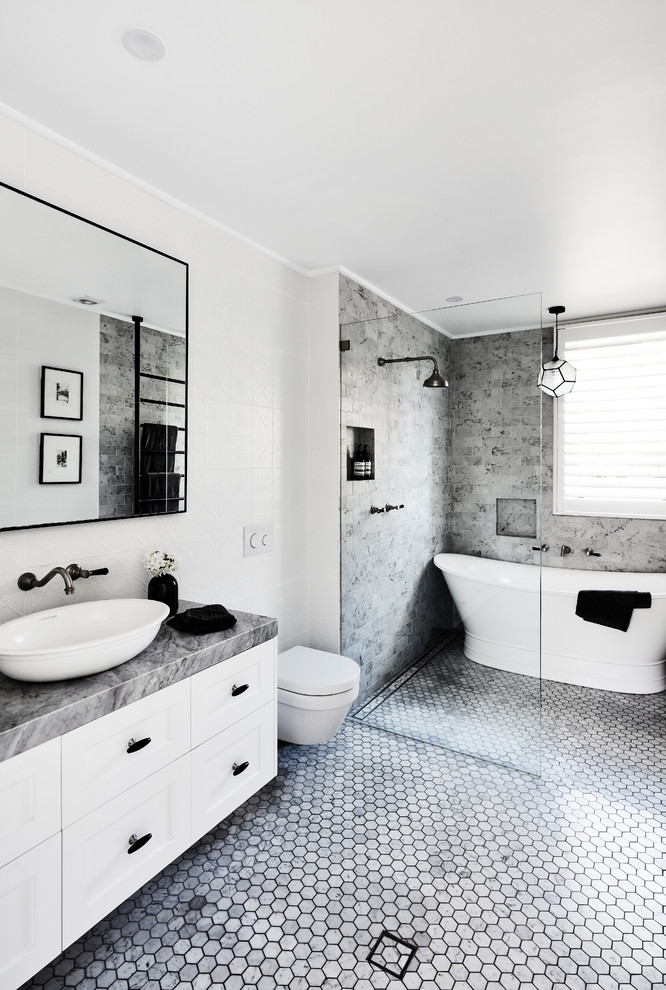 Inredning av ett klassiskt grå grått badrum med dusch, med skåp i shakerstil, vita skåp, ett fristående badkar, en kantlös dusch, en vägghängd toalettstol, grå kakel, vita väggar, ett fristående handfat och grått golv