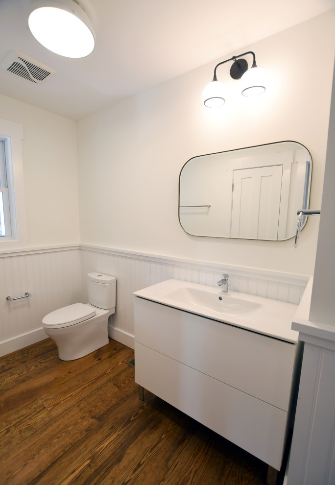 Bathroom - mid-sized cottage 3/4 medium tone wood floor, brown floor, single-sink and wainscoting bathroom idea in New York with flat-panel cabinets, white cabinets, a one-piece toilet, white walls, an integrated sink, solid surface countertops, white countertops and a freestanding vanity