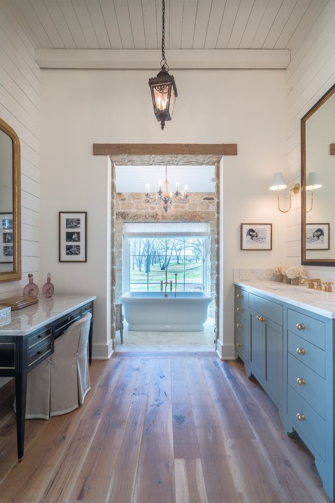Cette image montre une salle de bain rustique avec une baignoire indépendante, un sol en bois brun, un placard avec porte à panneau encastré, des portes de placard bleues, un mur blanc, un lavabo encastré, un sol marron et un plan de toilette blanc.