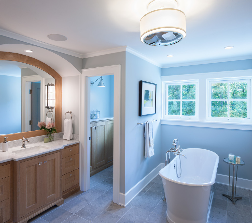 Cette image montre une grande salle de bain principale rustique en bois clair avec un placard à porte shaker, une baignoire indépendante, un mur bleu, un sol en carrelage de céramique, un lavabo encastré, un plan de toilette en marbre, un sol gris et un plan de toilette blanc.