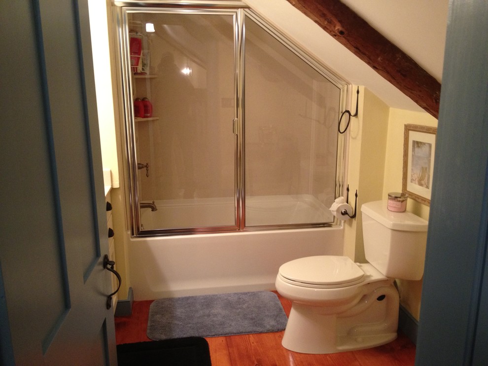 Inredning av ett lantligt litet badrum med dusch, med släta luckor, vita skåp, ett badkar i en alkov, en dusch/badkar-kombination, en toalettstol med separat cisternkåpa, gula väggar, mellanmörkt trägolv, ett undermonterad handfat och bänkskiva i akrylsten