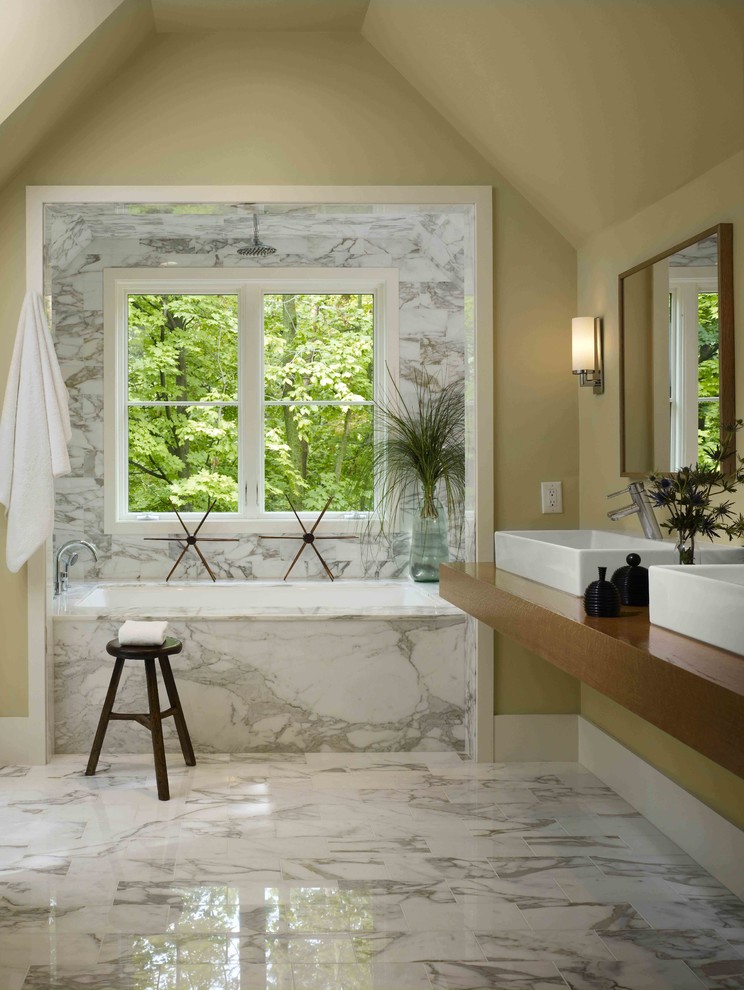 Lantlig inredning av ett badrum, med ett fristående handfat, träbänkskiva, ett undermonterat badkar, vit kakel, beige väggar och marmorgolv