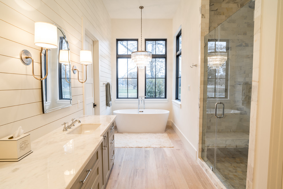 Bild på ett lantligt badrum, med luckor med infälld panel, grå skåp, ett fristående badkar, en dusch i en alkov, vita väggar, ett undermonterad handfat, beiget golv och dusch med gångjärnsdörr