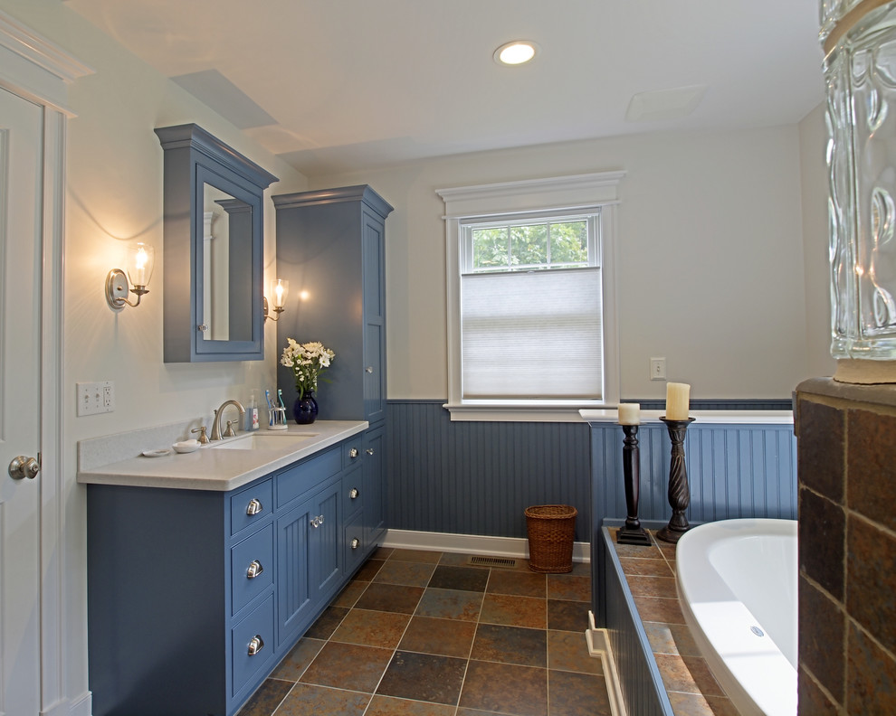 Cette image montre une salle de bain craftsman de taille moyenne avec un placard à porte shaker, des portes de placard bleues, une baignoire posée, un mur blanc, un sol en ardoise, un lavabo encastré et un plan de toilette en surface solide.