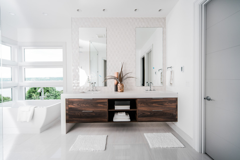 Идея дизайна: ванная комната в современном стиле с плоскими фасадами, темными деревянными фасадами, отдельно стоящей ванной, белой плиткой, белыми стенами, врезной раковиной, серым полом, белой столешницей и зеркалом с подсветкой