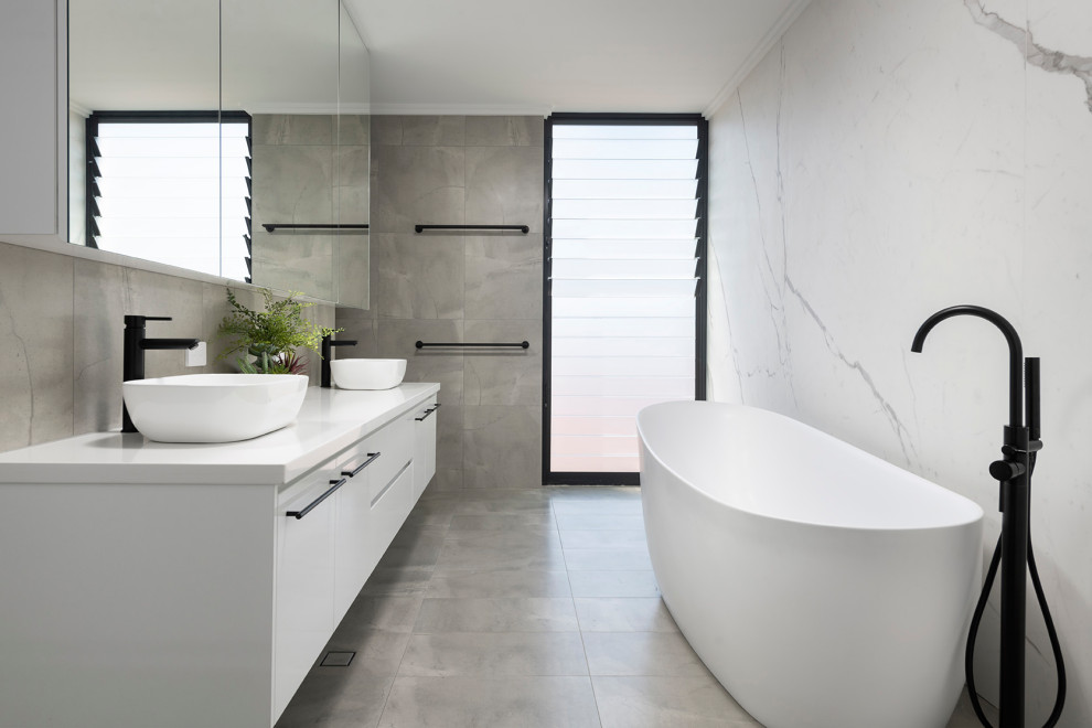 Bild på ett mellanstort funkis vit vitt en-suite badrum, med släta luckor, vita skåp, ett fristående badkar, grå kakel, porslinskakel, klinkergolv i småsten, ett fristående handfat och grått golv