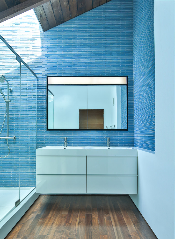 Inredning av ett modernt badrum med dusch, med släta luckor, vita skåp, en dusch i en alkov, blå kakel, stickkakel, mörkt trägolv och ett integrerad handfat