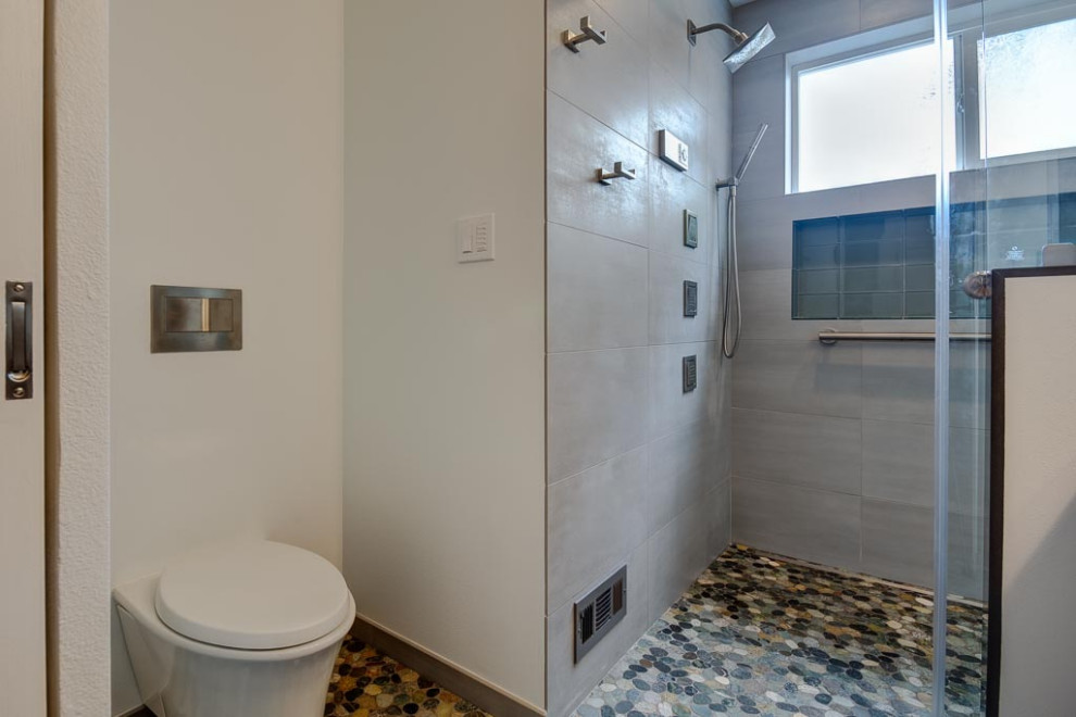 Bild på ett litet funkis grå grått badrum med dusch, med en kantlös dusch, en vägghängd toalettstol, grå kakel, vita väggar, klinkergolv i småsten, flerfärgat golv och med dusch som är öppen