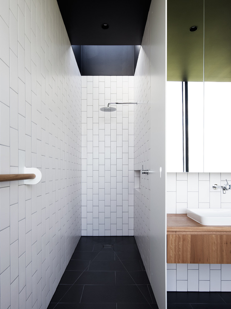 Пример оригинального дизайна: ванная комната в современном стиле с плоскими фасадами, фасадами цвета дерева среднего тона, душем без бортиков, белой плиткой, плиткой кабанчик, душевой кабиной, накладной раковиной, столешницей из дерева, черным полом и открытым душем