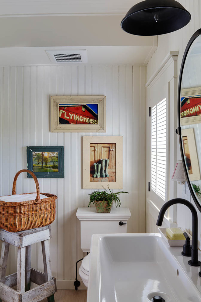Modelo de cuarto de baño marinero de tamaño medio con armarios con paneles con relieve, puertas de armario de madera clara, sanitario de una pieza, paredes blancas, suelo de madera clara, aseo y ducha y lavabo con pedestal