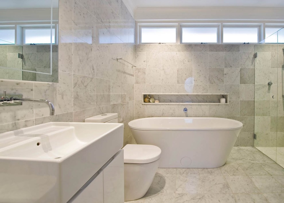 На фото: большая главная ванная комната в современном стиле с плоскими фасадами, белыми фасадами, отдельно стоящей ванной, душем в нише, белой плиткой, каменной плиткой, белыми стенами, мраморным полом, раковиной с несколькими смесителями, унитазом-моноблоком, серым полом и душем с распашными дверями с