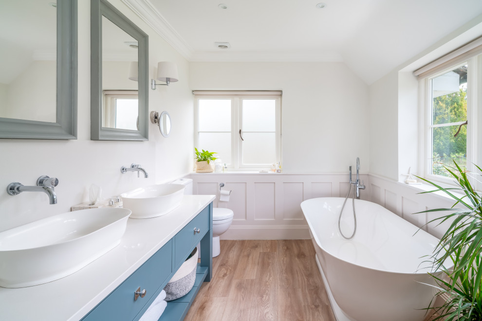 Design ideas for a traditional bathroom in Buckinghamshire with flat-panel cabinets, blue cabinets, a freestanding bath, white walls, medium hardwood flooring, a vessel sink, brown floors, white worktops, double sinks, a freestanding vanity unit, wainscoting and a dado rail.