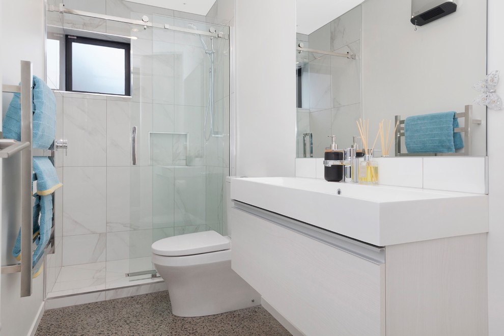 This is an example of a small contemporary family bathroom in Christchurch with a walk-in shower, a wall mounted toilet, white tiles, ceramic tiles, white walls, concrete flooring, an integrated sink and a sliding door.