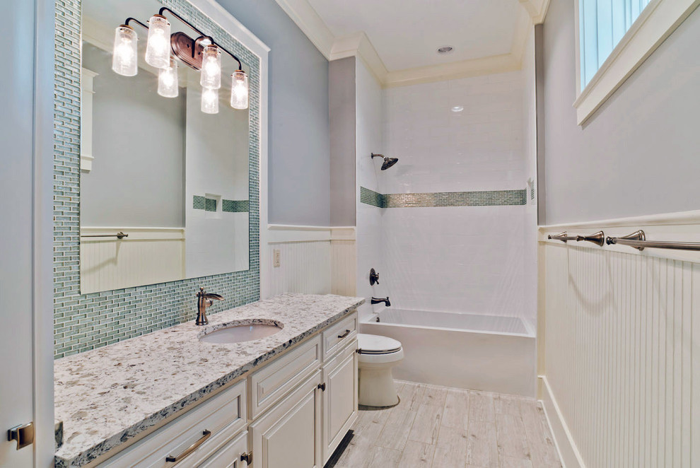 Foto på ett mellanstort maritimt badrum, med luckor med infälld panel, vita skåp, ett platsbyggt badkar, en dusch/badkar-kombination, en vägghängd toalettstol, grå kakel, stickkakel, grå väggar, ljust trägolv, ett undermonterad handfat och granitbänkskiva