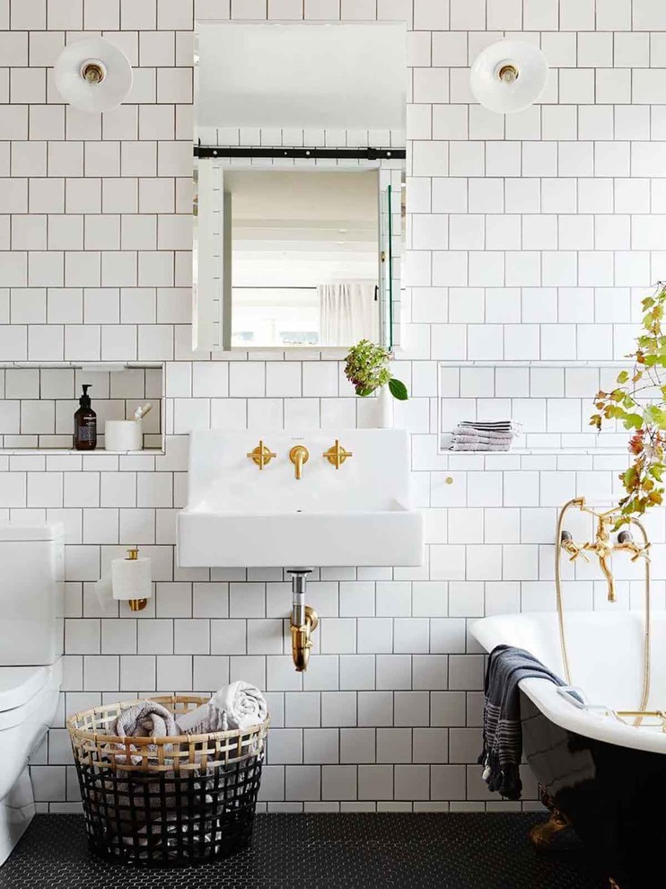 Foto på ett vintage badrum med dusch, med ett badkar med tassar, en toalettstol med separat cisternkåpa, vit kakel, ett väggmonterat handfat och svart golv