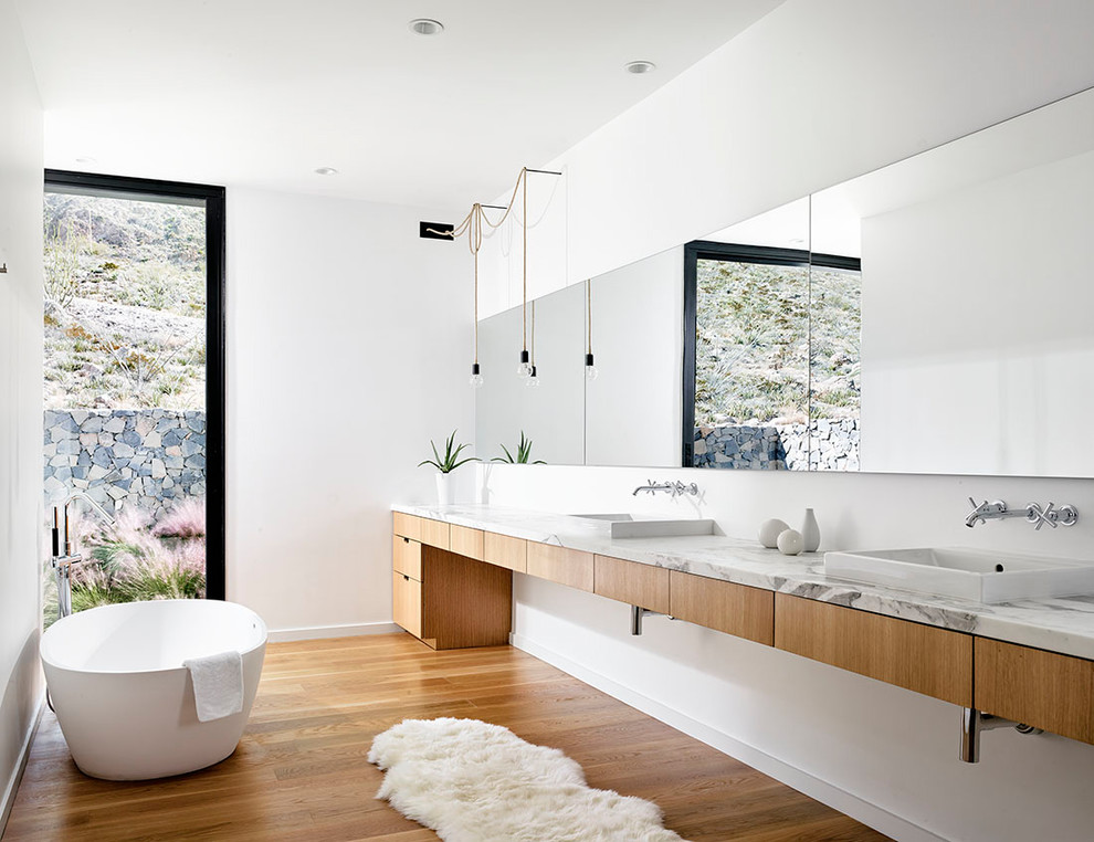 Inspiration for a modern ensuite bathroom in Austin with flat-panel cabinets, brown cabinets, a freestanding bath, white walls, medium hardwood flooring, a built-in sink, marble worktops and beige floors.