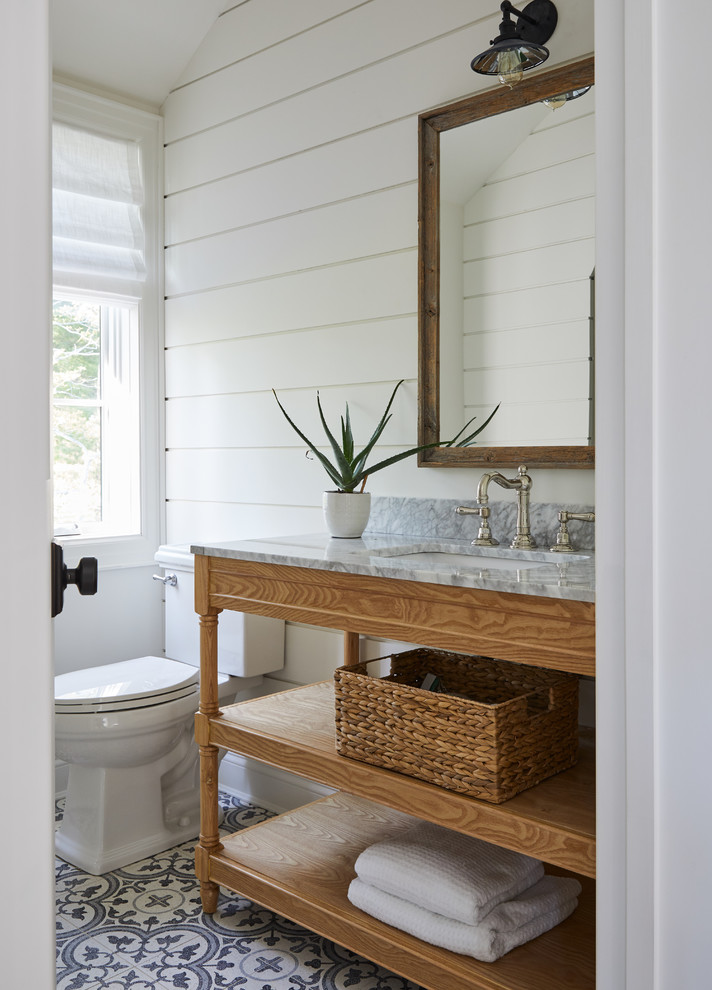 Lantlig inredning av ett grå grått badrum med dusch, med öppna hyllor, skåp i mellenmörkt trä, en toalettstol med separat cisternkåpa, vita väggar, cementgolv, ett undermonterad handfat och flerfärgat golv