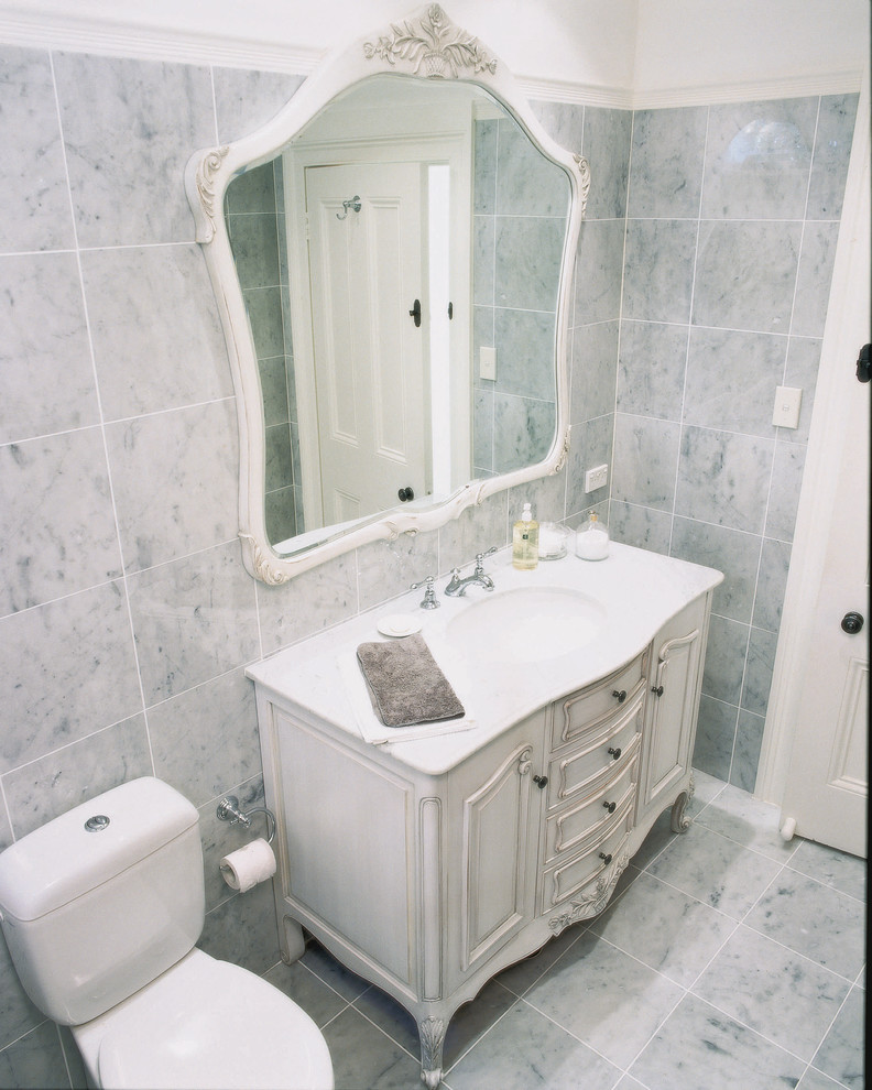 Modelo de cuarto de baño tradicional de tamaño medio con puertas de armario blancas, bañera encastrada sin remate, sanitario de dos piezas, baldosas y/o azulejos grises, paredes grises, lavabo bajoencimera, encimera de mármol y suelo gris