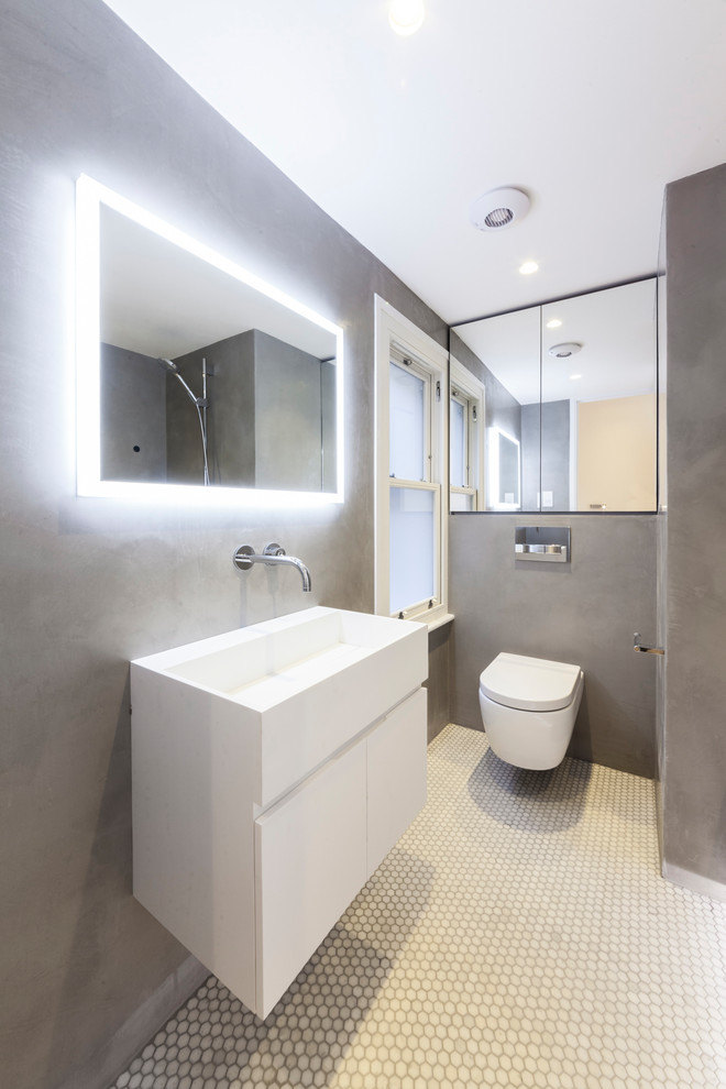 Bild på ett funkis badrum, med släta luckor, vita skåp, en vägghängd toalettstol, grå väggar, ett väggmonterat handfat och vitt golv