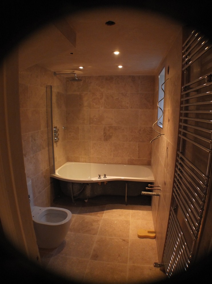 Photo of a small traditional family bathroom in London with a built-in bath, a shower/bath combination, flat-panel cabinets, medium wood cabinets, a two-piece toilet, beige tiles, travertine tiles, beige walls, travertine flooring, a console sink, beige floors and a hinged door.