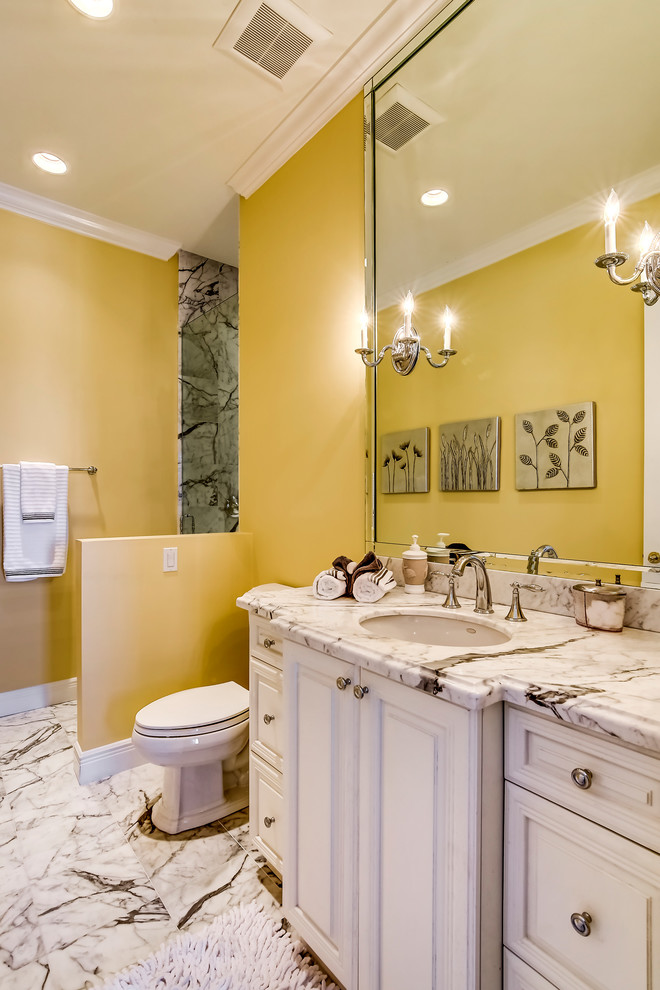 Cette image montre une salle d'eau traditionnelle de taille moyenne avec un lavabo posé, un placard avec porte à panneau surélevé, des portes de placard blanches, un plan de toilette en marbre, une douche d'angle, WC séparés, un carrelage blanc, des dalles de pierre, un mur jaune et un sol en marbre.