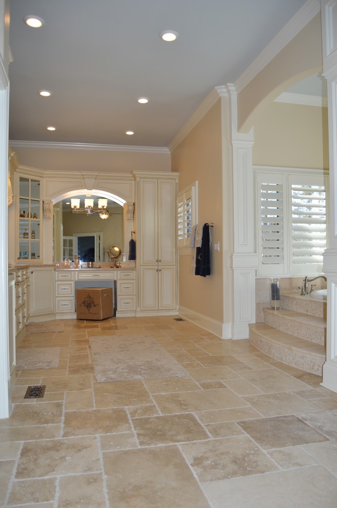 Пример оригинального дизайна: большая главная ванная комната в стиле неоклассика (современная классика) с врезной раковиной, фасадами с выступающей филенкой, белыми фасадами, столешницей из известняка, накладной ванной, двойным душем, унитазом-моноблоком, бежевой плиткой, каменной плиткой, бежевыми стенами и полом из известняка
