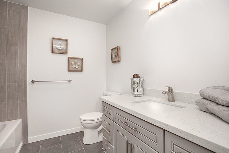 Cette image montre une salle de bain traditionnelle avec un placard avec porte à panneau surélevé, des portes de placard grises, une baignoire en alcôve, un combiné douche/baignoire, WC à poser, un carrelage gris, des carreaux de porcelaine, un mur blanc, un sol en carrelage de porcelaine, un lavabo encastré et un plan de toilette en quartz.