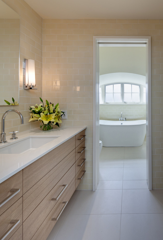 Cette image montre une salle de bain principale design en bois clair de taille moyenne avec un placard à porte plane, une baignoire indépendante, un carrelage beige, des carreaux de céramique, un mur beige, un sol en carrelage de porcelaine, un lavabo encastré et un plan de toilette en quartz modifié.